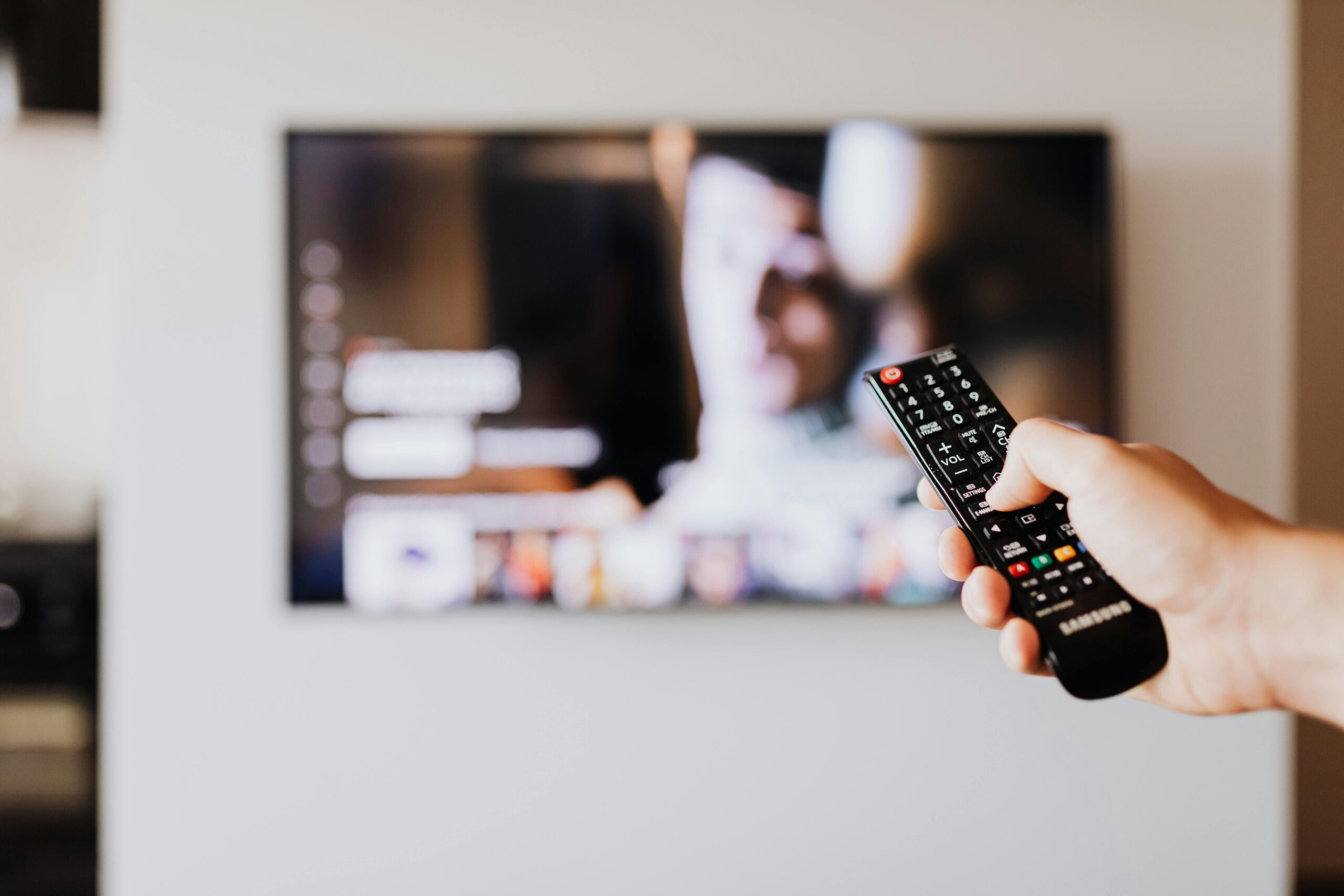 Home Person using a TV remote control with a blurred television screen in the background.